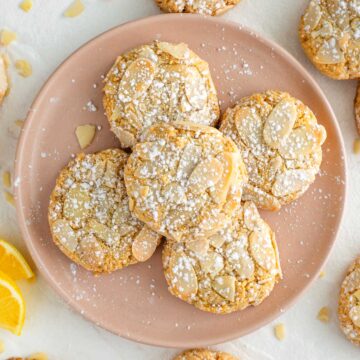 Top down view of almond tahini cookies on a pink plate. The cookies are lightly browned around the edges, topped with sliced almonds, and dusted with powdered sugar.